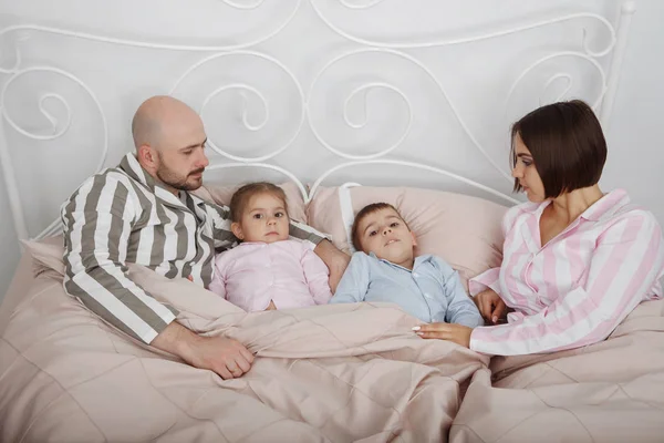 Young pretty woman mother and bald man father with son and daughter in pajamas lie in bed with pillows and pink bedclothes.