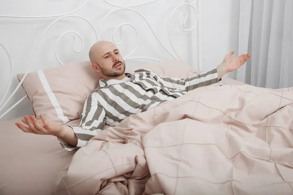 Handsome bald, bearded man in striped pajamas is lying and thinking in bed with pillows and pink bedding.