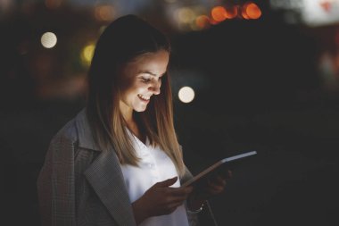 Güzel genç kadın bir tablet vasıl çok renkli ışıklar arka plan üzerinde akşam şehrin sokakta arıyor. Yüzü cihazın ışıkla aydınlatılmış.