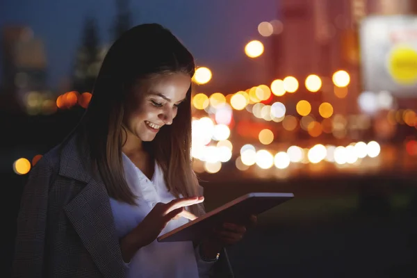 美しい若い女性は マルチカラーのライトの背景に夕方の都市の路上でタブレットを見ています デバイスの光に顔を照らされました — ストック写真