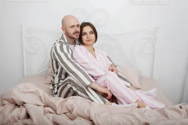 Young pretty woman and a bald bearded man in pajamas hug and caress in bed with pillows and pink bedclothes.