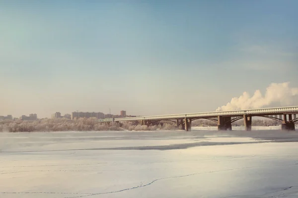 Coast of the winter river in Siberia. Skyline with outlines of houses and steam above the water. Bridge.