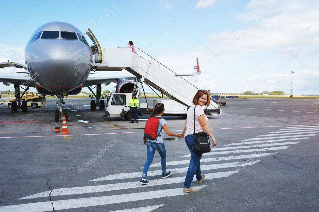 Mom with a bag and a daughter with a red backpack passengers go to the ramp of the plane on the airport field.