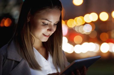 Güzel genç kadın bir tablet vasıl çok renkli ışıklar arka plan üzerinde akşam şehrin sokakta arıyor. Yüzü cihazın ışıkla aydınlatılmış.