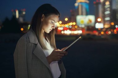 Güzel genç kadın bir tablet vasıl çok renkli ışıklar arka plan üzerinde akşam şehrin sokakta arıyor. Yüzü cihazın ışıkla aydınlatılmış.