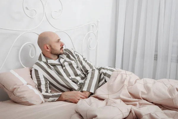 Handsome bald, bearded man in striped pajamas is lying and thinking in bed with pillows and pink bedding.