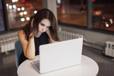 Masada laptop ile çalışma gece ofiste iş kadını. Pencerenin dışında çeşitli renklerde ışık yanıyor. Son Tarih, fazla mesai.