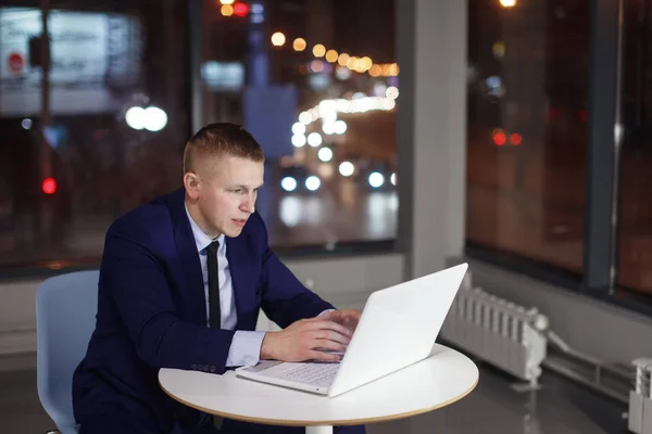 Businessman working at the table with laptop at night in the office. Outside the window are lit multicolored lights. Deadline, overtime.