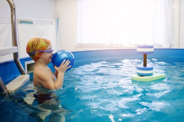 Çocuk bir sarı şapka ve gözlük bir topla oynamayı havuzda merdivenlerde oturuyor. Sağlıklı bir yaşam tarzı. Aquatherapy. Yüzme rehabilitasyon.