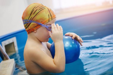 Çocuk bir sarı şapka ve gözlük bir topla oynamayı havuzda merdivenlerde oturuyor. Sağlıklı bir yaşam tarzı. Aquatherapy. Yüzme rehabilitasyon.
