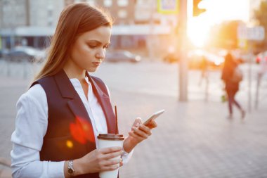 Genç güzel kadın ile smartphone ve kağıt kahve Kupası güneşin arka plan üzerinde şehir sokakta günbatımında. Telefona bakar. Vintage sesi filtre efekti renk stili.