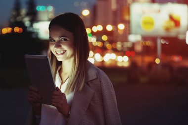 Güzel genç kadın bir tablet vasıl çok renkli ışıklar arka plan üzerinde akşam şehrin sokakta arıyor. Yüzü cihazın ışıkla aydınlatılmış.