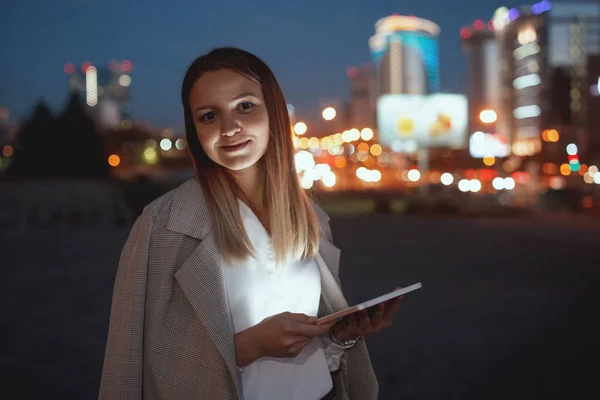 多色ライトの背景に夕方の都市の通りにタブレットと美しい若い女性 デバイスの光に顔を照らされました — ストック写真