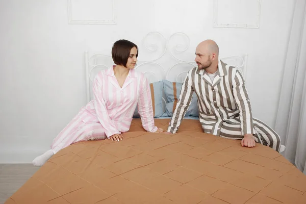 Young pretty woman and a bald bearded man in pajamas talk in bed with pillows and brown bedclothes.