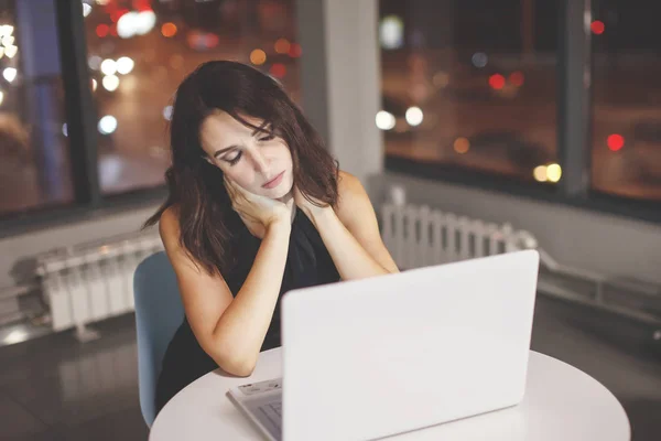 Businesswoman working at the table with laptop at night in the office. Outside the window are lit multicolored lights. Deadline, overtime.