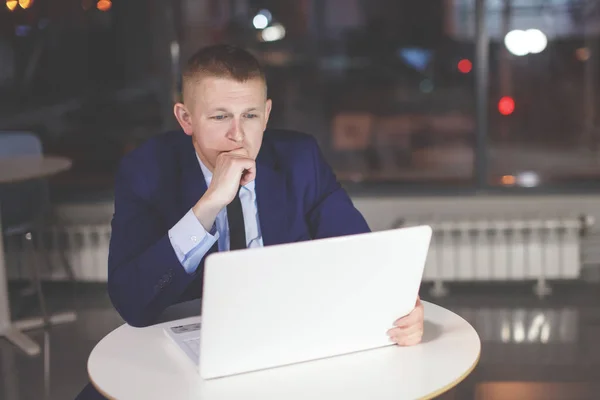 Businessman working at the table with laptop at night in the office. Outside the window are lit multicolored lights. Deadline, overtime.