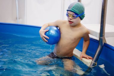 Çocuk bir sarı şapka ve gözlük bir topla oynamayı havuzda merdivenlerde oturuyor. Sağlıklı bir yaşam tarzı. Aquatherapy. Yüzme rehabilitasyon.