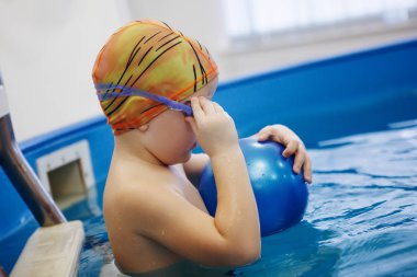 Çocuk bir sarı şapka ve gözlük bir topla oynamayı havuzda merdivenlerde oturuyor. Sağlıklı bir yaşam tarzı. Aquatherapy. Yüzme rehabilitasyon.