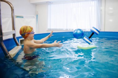 Çocuk bir sarı şapka ve gözlük bir topla oynamayı havuzda merdivenlerde oturuyor. Sağlıklı bir yaşam tarzı. Aquatherapy. Yüzme rehabilitasyon.