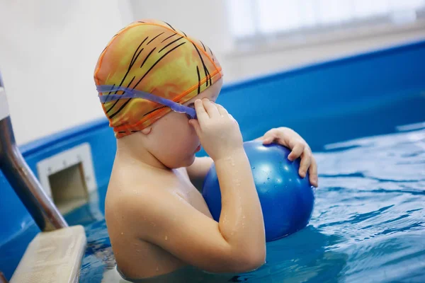 Çocuk bir sarı şapka ve gözlük bir topla oynamayı havuzda merdivenlerde oturuyor. Sağlıklı bir yaşam tarzı. Aquatherapy. Yüzme rehabilitasyon.