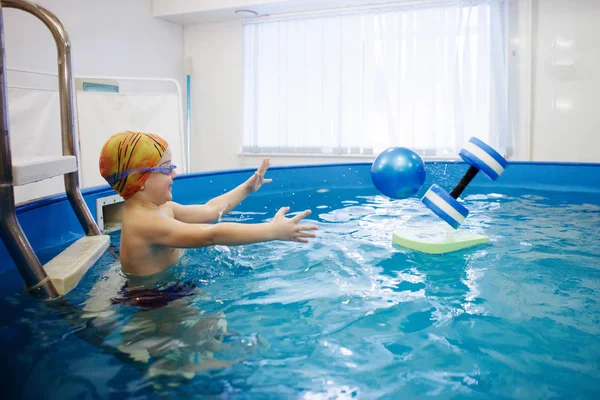 Çocuk bir sarı şapka ve gözlük bir topla oynamayı havuzda merdivenlerde oturuyor. Sağlıklı bir yaşam tarzı. Aquatherapy. Yüzme rehabilitasyon.