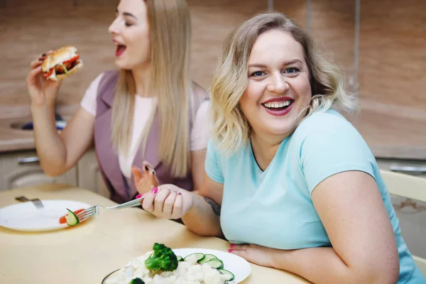 若いかなり脂肪ブロンドの女性は ジューシーなハンバーガーと笑いを食べるスリムなガール フレンドを羨ましきます 概念の健康的な食事 ダイエット 肥満と戦う — ストック写真