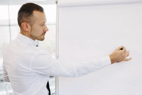 Teamwork of office staff. Businessman coach in a white shirt and tie, standing near the flipchart, explains the schedule of tasks for the future. Business training. Personal growth.