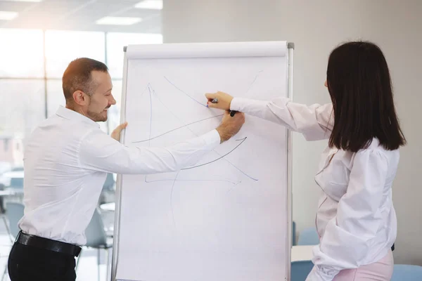 Teamwork of office staff. Businessman coach in a white shirt and tie, standing near a flipchart, explains to the woman the schedule of tasks for the future. Business training. Personal growth.