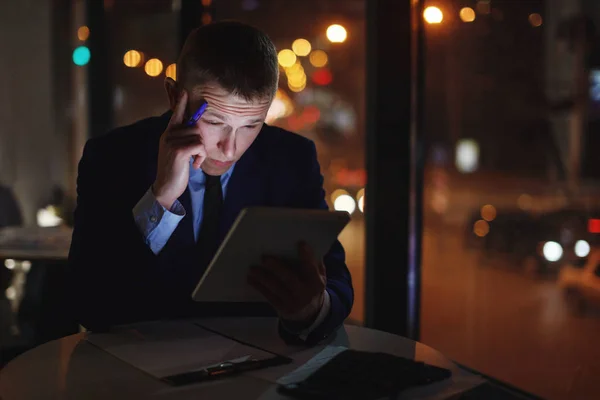 Office manager sits at a table at night, looks at a digital tablet with a touchscreen and solves an urgent problem.