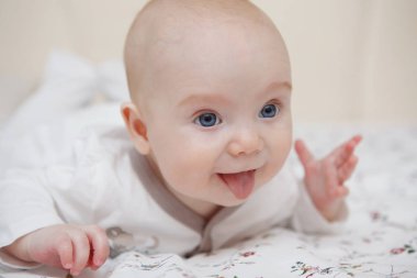 Güzel beş aylık mavi gözlü kız bebek karnında yatıyor. Gösterir dil, çeşitli duygu ve yüzünde bir gülümseme. Bebek bak. Anne bakım. Çocuk Bakımı. Yakın çekim.
