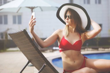 Genç kadın bir kırmızı mayo güzel ve mavi su resort otel ile yapay rattan havuz kenarında şezlong içinde oturan smartphone içinde plaj şapka görünüyor.