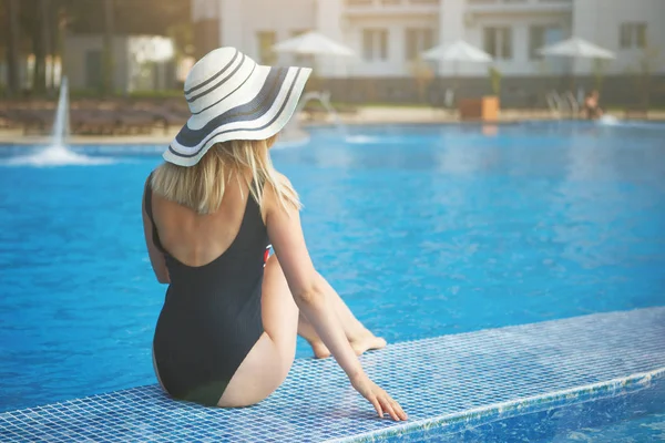 Uzun güzel bacaklar ile plaj şapkalı bir kadın mavi su ile bir havuz kenarına oturur. Arkadan görünüm.