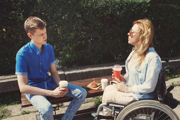 Woman in wheelchair with boyfriend drinking coffee and eating muffins in city summer park