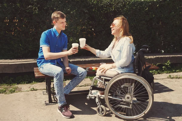Woman in wheelchair with boyfriend drinking coffee and eating muffins in city summer park