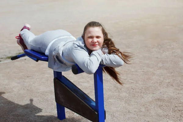 A young teenage girl in a gray sports suit, with long hair smiling and doing outdoor exercises on street exercisers.