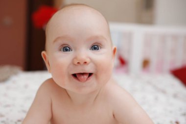 Güzel beş aylık mavi gözlü kız bebek karnında yatıyor. Gösterir dil, çeşitli duygu ve yüzünde bir gülümseme. Bebek bak. Anne bakım. Çocuk Bakımı. Yakın çekim.