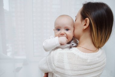 Genç mutlu anne, bebeğini kollarında pencereye doğru öpüyor. Annelik bakımı. Çocuk bakımı. Yakın plan..