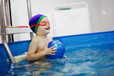 Mavi şapkalı ve gözlüklü çocuk havuzda topla oynuyor..