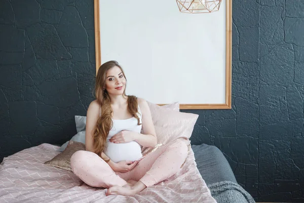 Lykkelig, gravid kvinne som sitter i sengen hjemme – stockfoto