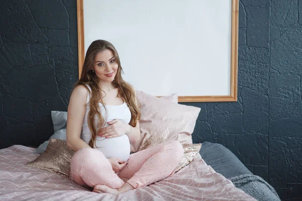 Lykkelig, gravid kvinne som sitter i sengen hjemme – stockfoto