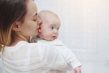 Genç mutlu anne, bebeğini kollarında pencereye doğru öpüyor. Annelik bakımı. Çocuk bakımı. Yakın plan..