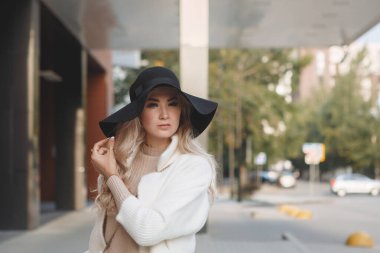 Portrait of a blonde lady in a black hat outdoors, long hair, beige coat.