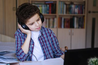Kulaklıklı genç çocuk düşünceli bir şekilde müzik dinliyor, masada oturuyor. Evde koronavirüs karantinası eğitimi alıyorum. Seçici odaklanma. Bulanık arkaplan.