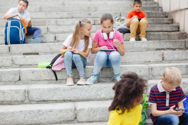 Farklı milletlerden bir grup öğrenci, renkli giysiler içinde taş basamaklarda oturuyorlar. Gençler konuşuyor, kulaklıkla müzik dinliyor, kitap okuyor..