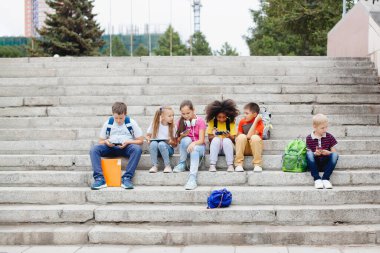 Farklı milletlerden bir grup öğrenci, renkli giysiler içinde taş basamaklarda oturuyorlar. Gençler konuşuyor, kulaklıkla müzik dinliyor, kitap okuyor..