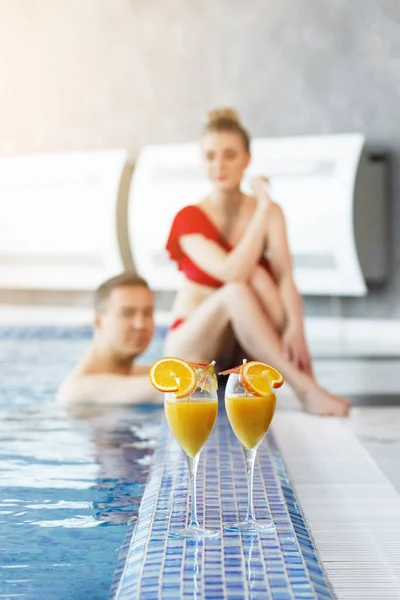 两杯橙汁和切片放在游泳池里的一对夫妇的背景上 穿着红色游泳衣的女人坐在边缘 男人在蓝色的水里游泳 有选择的重点 背景模糊 垂直方向 — 图库照片