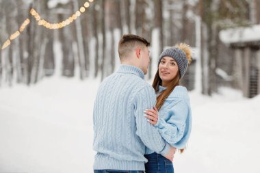 Kışlık bir parkta kucaklaşan sevimli bir çift. Sıcak kıyafetler, örgü şapkalar, kar yığınları, ağaçlar, bulanık arka plan..