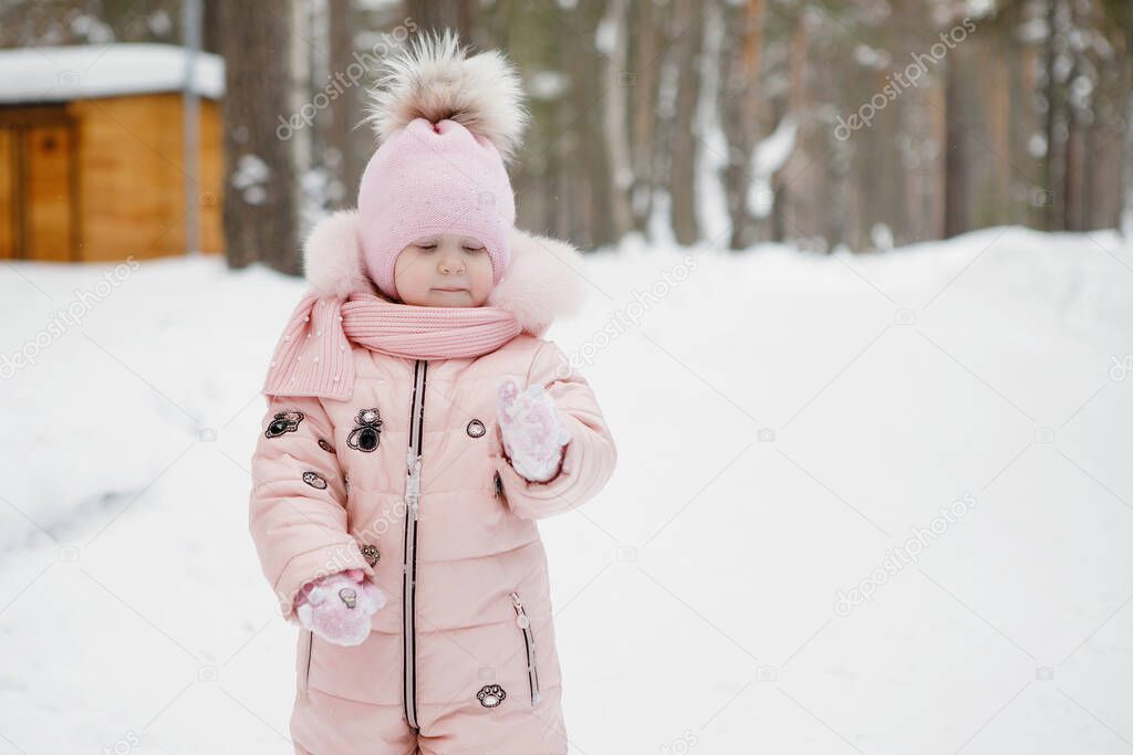 Girl baby in pink overalls in a cap with a pompon is walking in the white snow. Sunny winter day in a city park, outdoors.