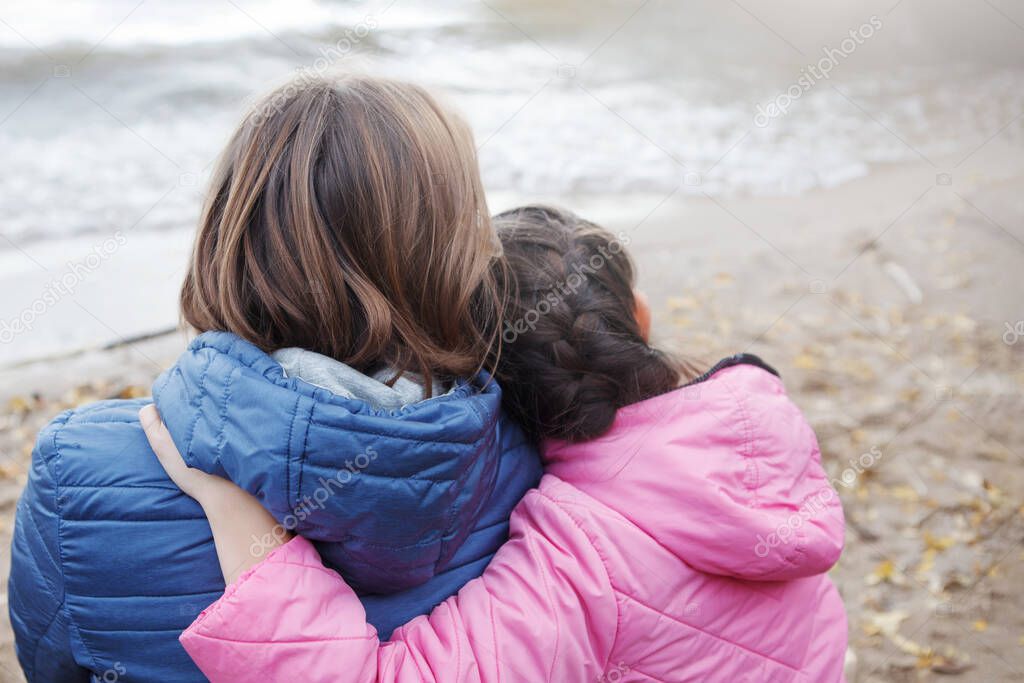 Mom and daughter sit hugging on the shore. Pink and blue jackets, long hair. Family happiness. Back view. Selective focus, blur background.