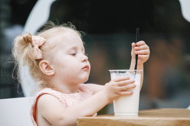 Sarışın ve milkshake, şehirdeki bir masada. Pembe elbiseli, at kuyruklu üç yaşında bir bebek kameraya duygusal pozlar veriyor..